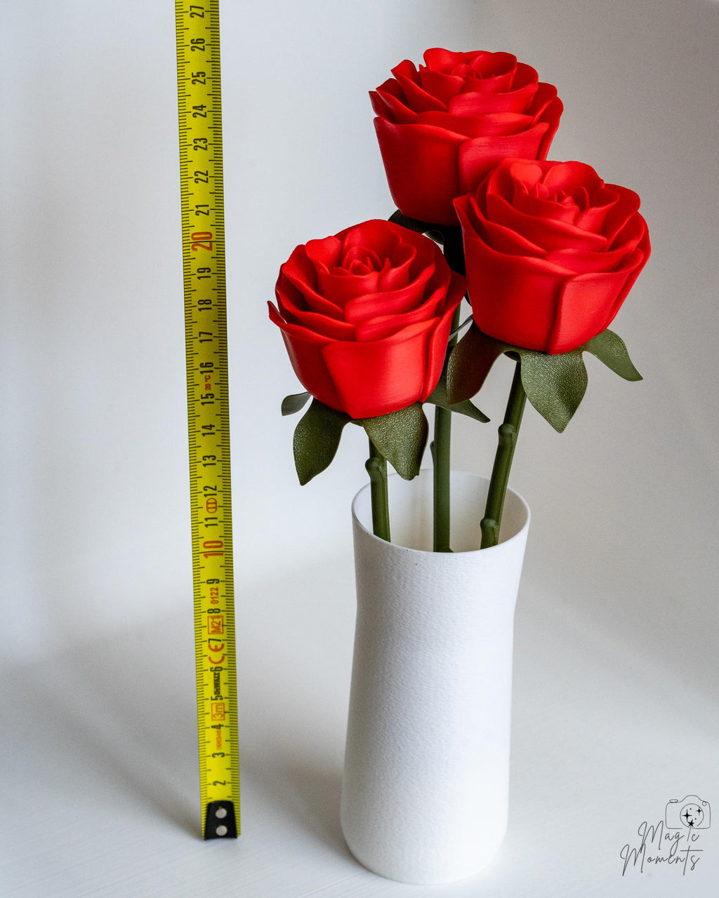 Three red roses in a white vase with a measuring tape on a light gray background