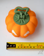 Orange pumpkin-shaped container with green lid on a white background, measured by a tape measure.