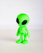 A small green alien figurine with large black eyes, standing against a plain white background.