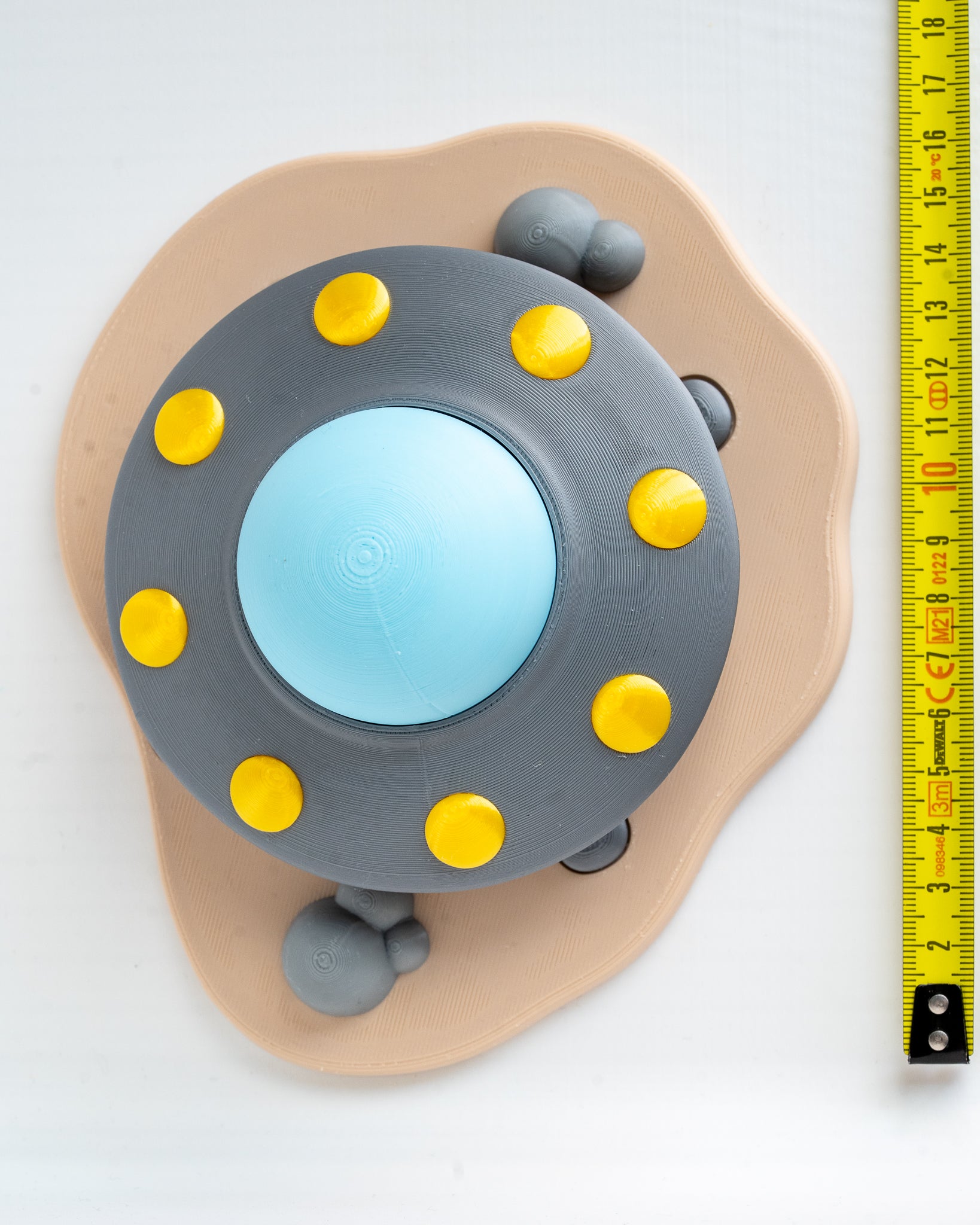 A toy or model shaped like a round gray UFO with a light blue center, yellow circular details, and a beige irregular base, shown on a white background with a yellow measuring tape along the right edge.