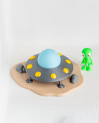 A toy UFO spacecraft with a light blue dome, gray body, yellow circular details, and a beige landing pad, shown on a white background next to a small green alien figurine.
