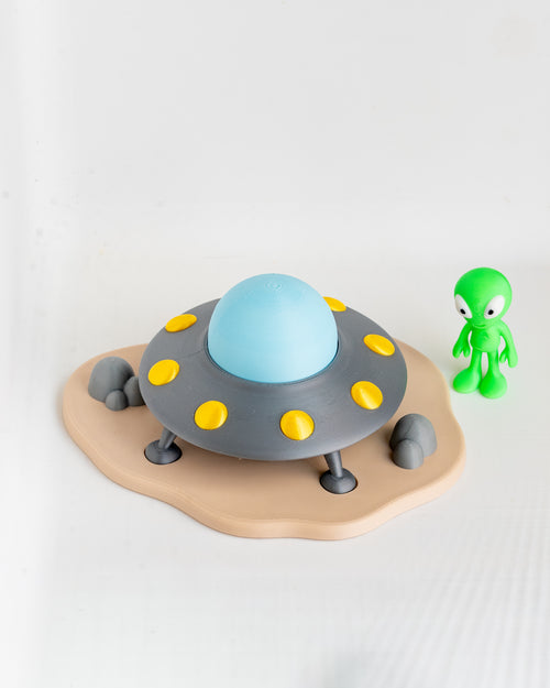 A toy UFO spacecraft with a light blue dome, gray body, yellow circular details, and a beige landing pad, shown on a white background next to a small green alien figurine.