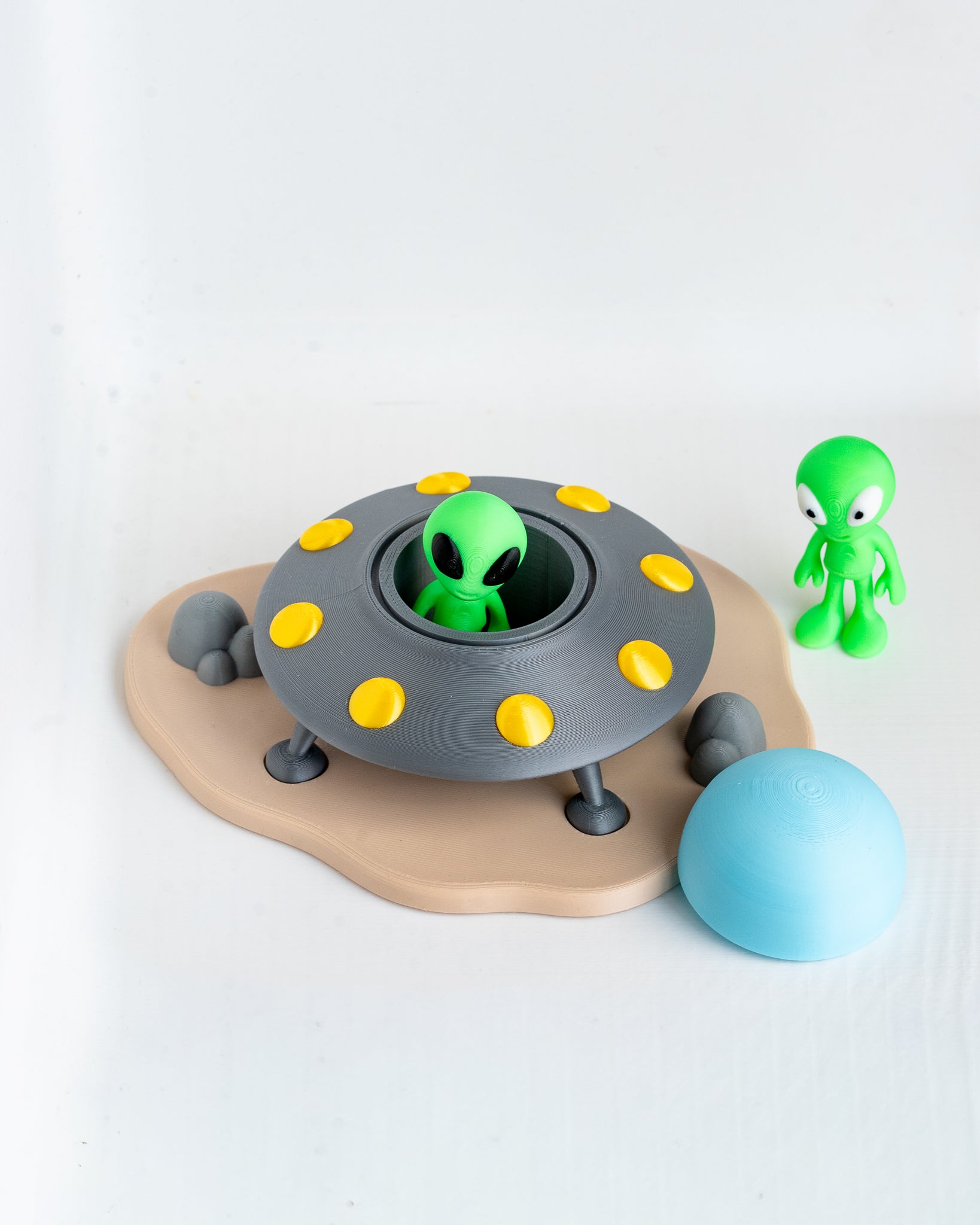 A children's toy set featuring a gray flying saucer with yellow lights, a green alien sitting inside, a small green alien figure standing beside it, a light blue oval ball, and a tan base on a white background.