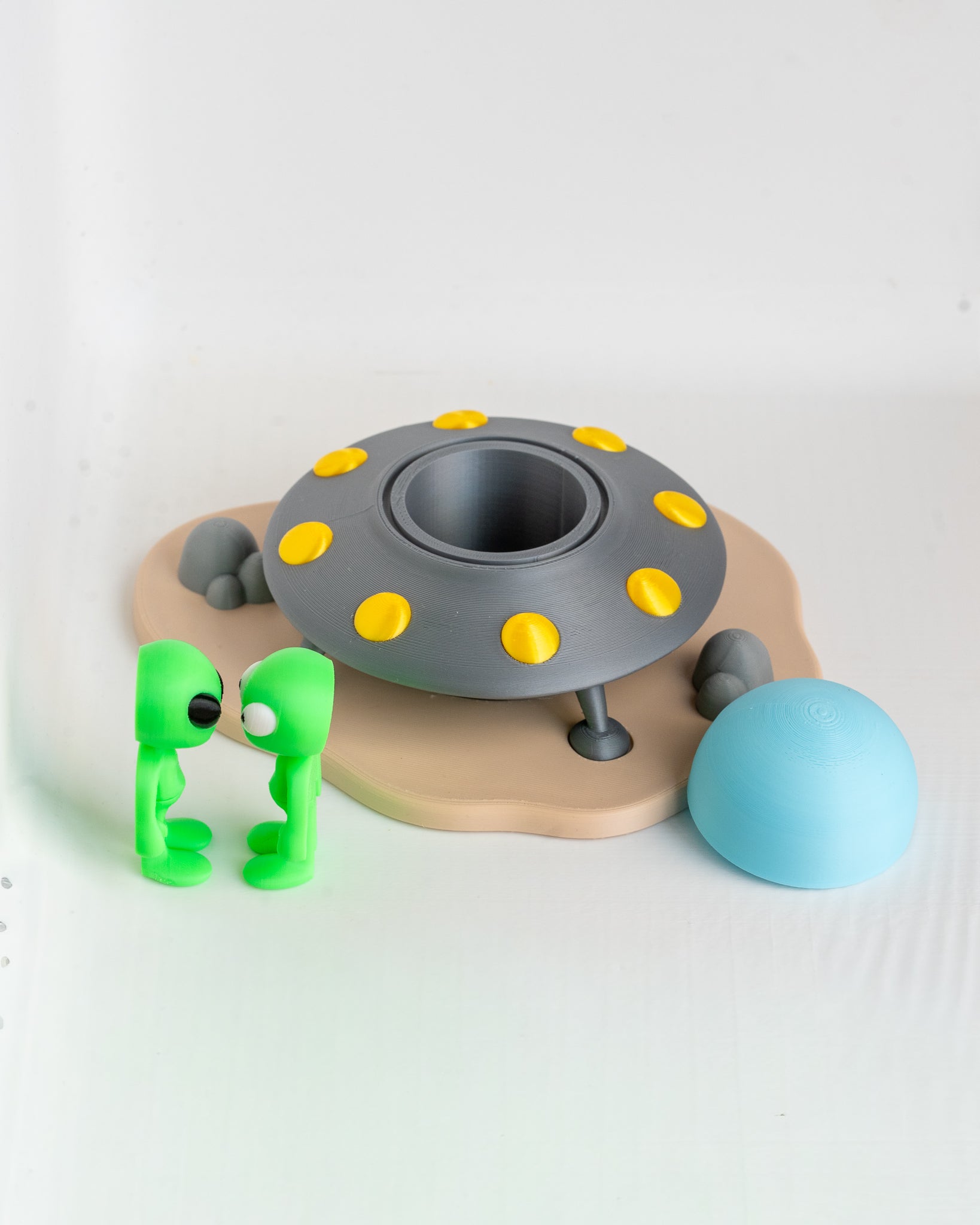 A toy model featuring a gray flying saucer with yellow accents, a beige landing pad, two small green alien figures, and a light blue sphere on a plain white background.