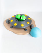 A toy playset featuring a gray flying saucer with yellow lights, two green cartoon aliens peeking out, a tan cloud-shaped base with small gray rocks, and a light blue ball on a white background.