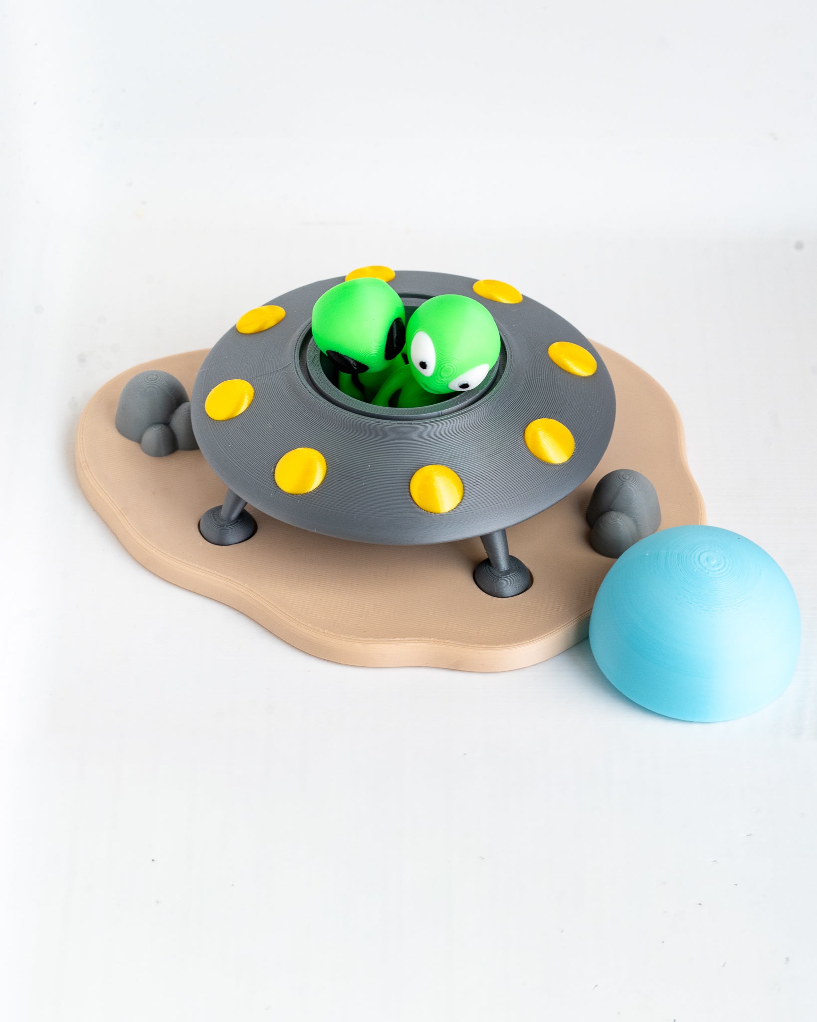 A toy playset featuring a gray flying saucer with yellow lights, two green cartoon aliens peeking out, a tan cloud-shaped base with small gray rocks, and a light blue ball on a white background.