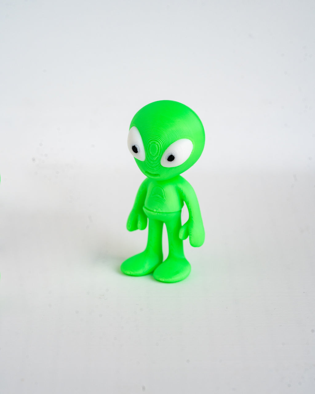 A small green alien figurine with large white eyes, standing on a plain light gray background.