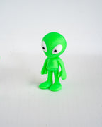 A small green alien figurine with large white eyes, standing on a plain light gray background.