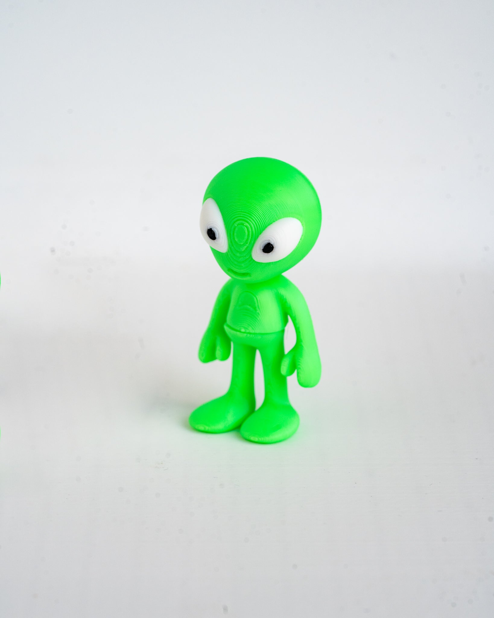 A small green alien figurine with large white eyes, standing on a plain light gray background.