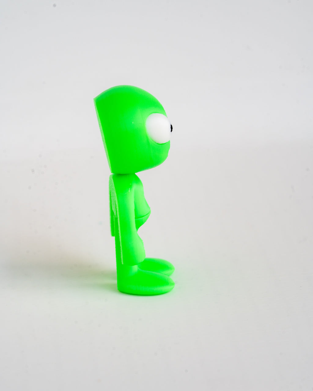 A small bright green alien figurine with a large white eye and black pupil, standing in profile against a plain light gray background.
