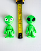 Two small green alien figurines shown on a white background with a yellow tape measure between them for scale.