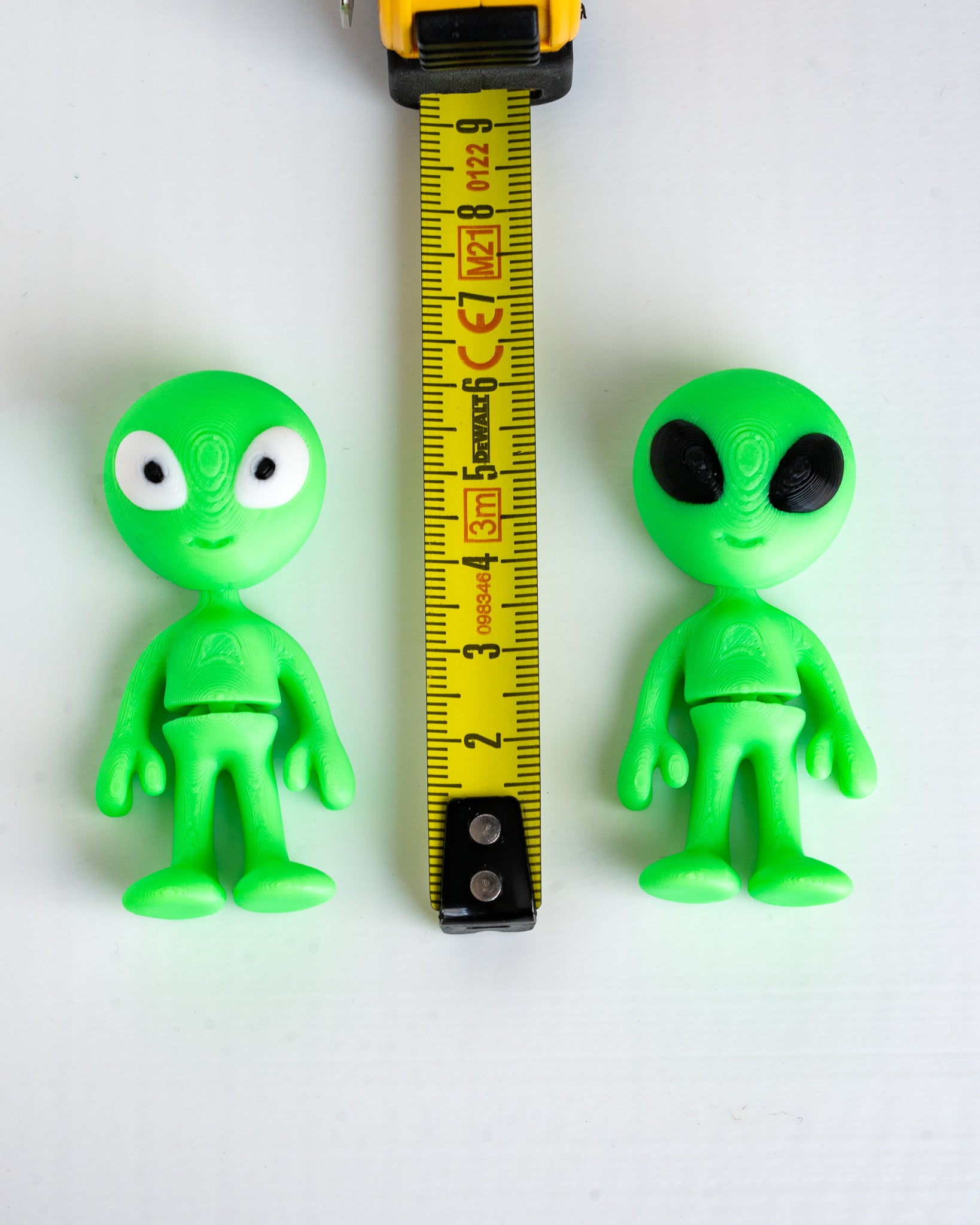 Two small green alien figurines shown on a white background with a yellow tape measure between them for scale.