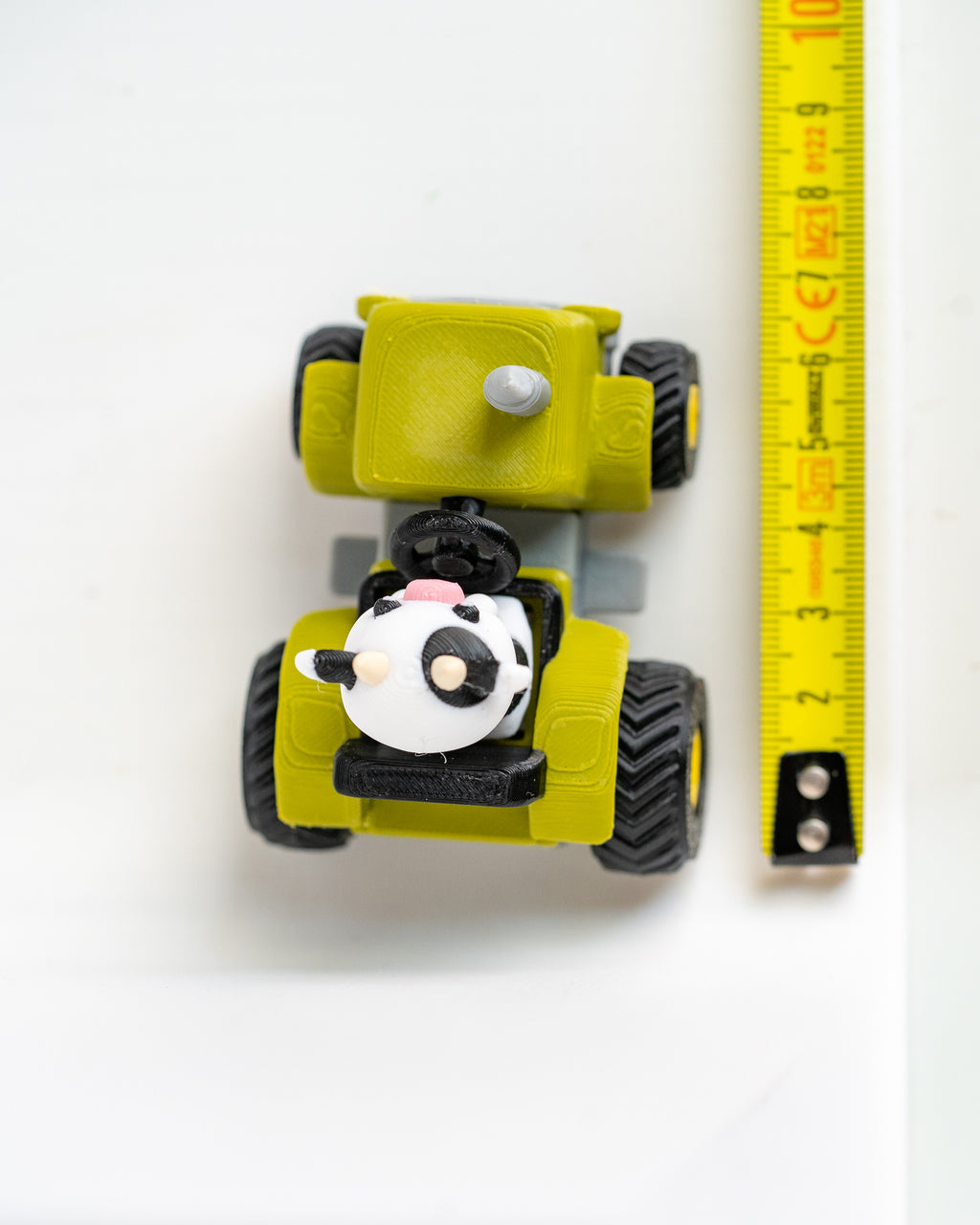 A small yellow toy construction vehicle with black wheels and a cow-shaped driver, shown next to a yellow measuring tape on a white background.