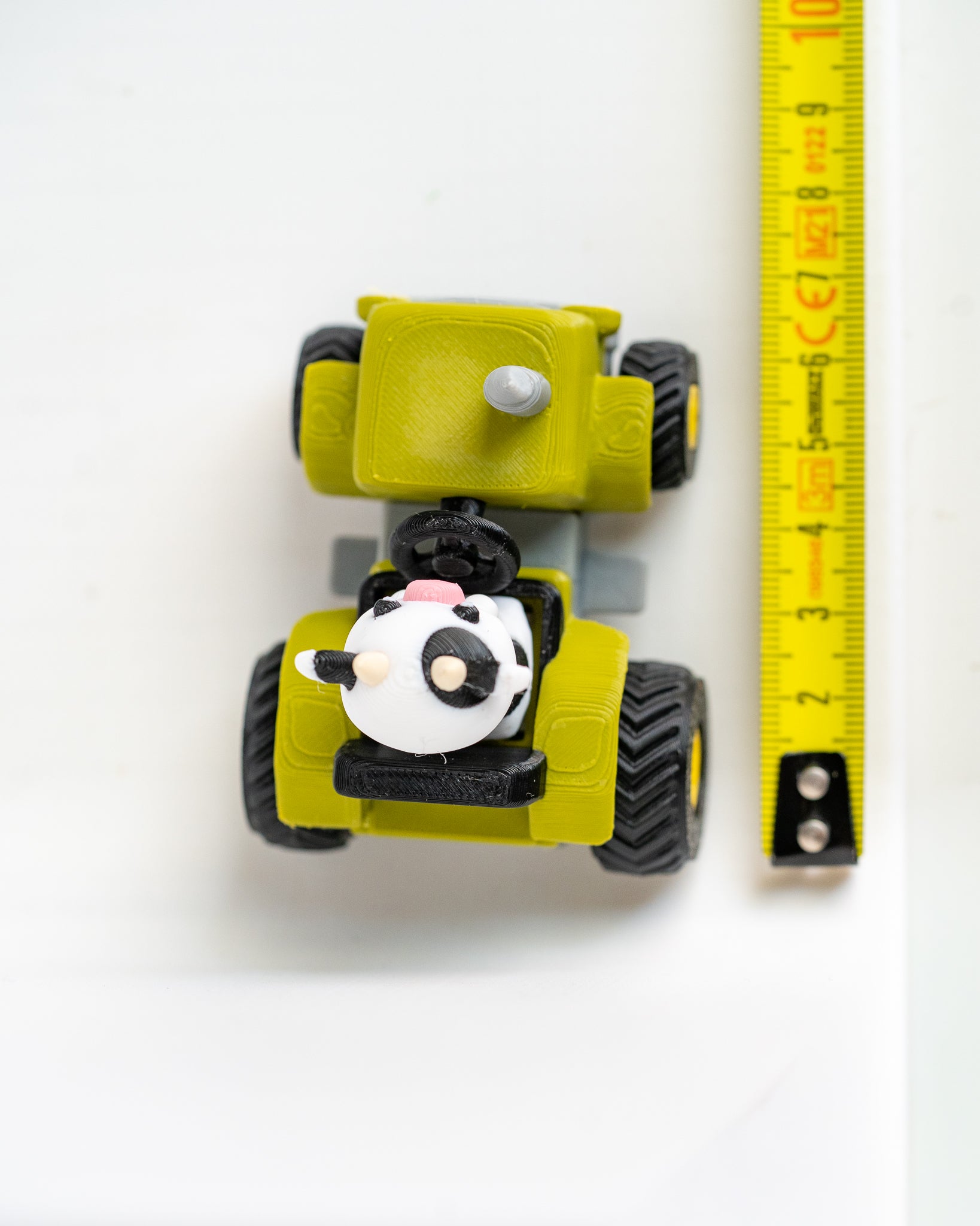 A small yellow toy construction vehicle with black wheels and a cow-shaped driver, shown next to a yellow measuring tape on a white background.