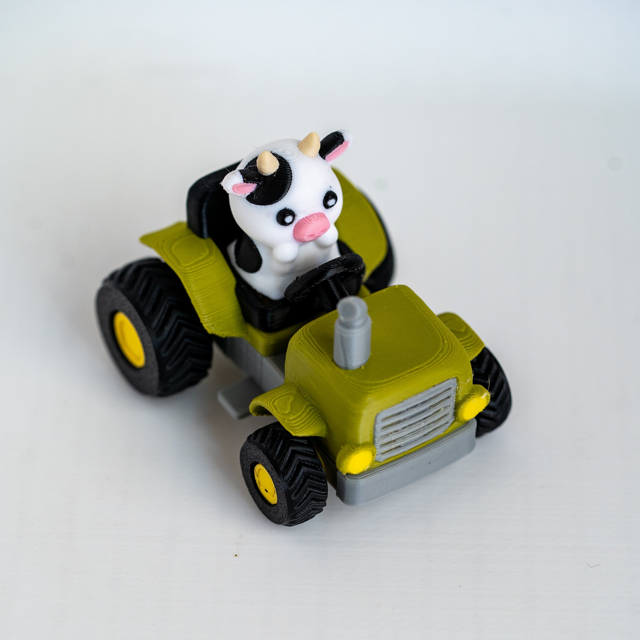 A small toy tractor in olive green and black with yellow wheels, featuring a cute black-and-white cow figurine driving it, photographed on a plain light background.