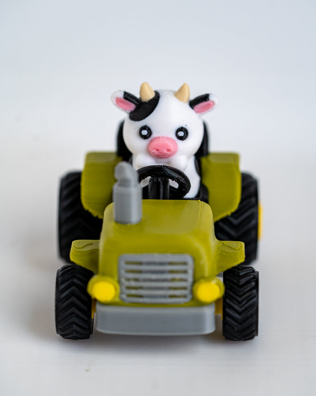 A small toy tractor in olive green and gray with black wheels, featuring a cute black-and-white cow character driving it, photographed on a plain light background.