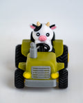 A small toy tractor in olive green and gray with black wheels, featuring a cute black-and-white cow character driving it, photographed on a plain light background.