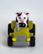 A small toy tractor in olive green and gray with black wheels, featuring a cute black-and-white cow character driving it, photographed on a plain light background.
