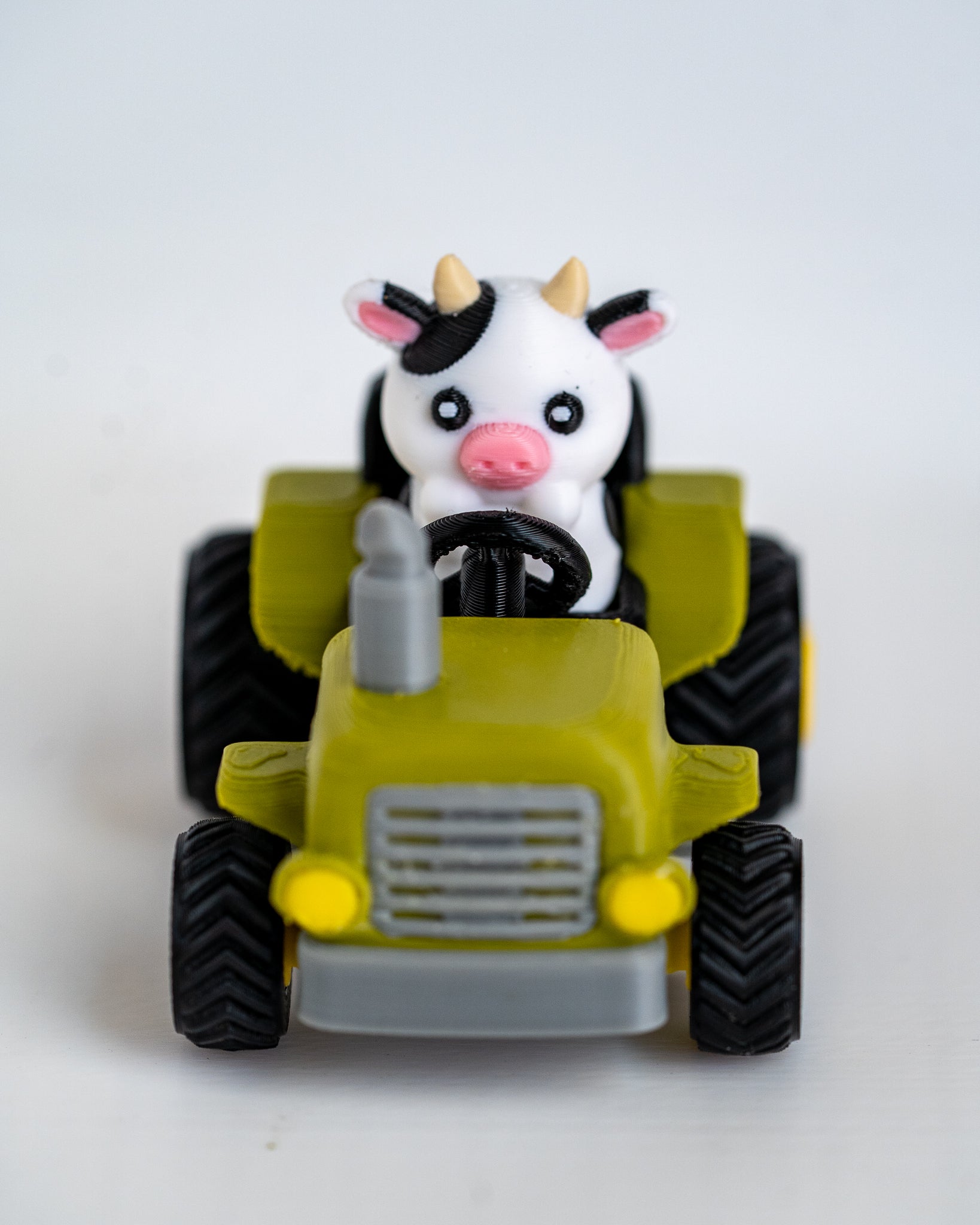 A small toy tractor in olive green and gray with black wheels, featuring a cute black-and-white cow character driving it, photographed on a plain light background.