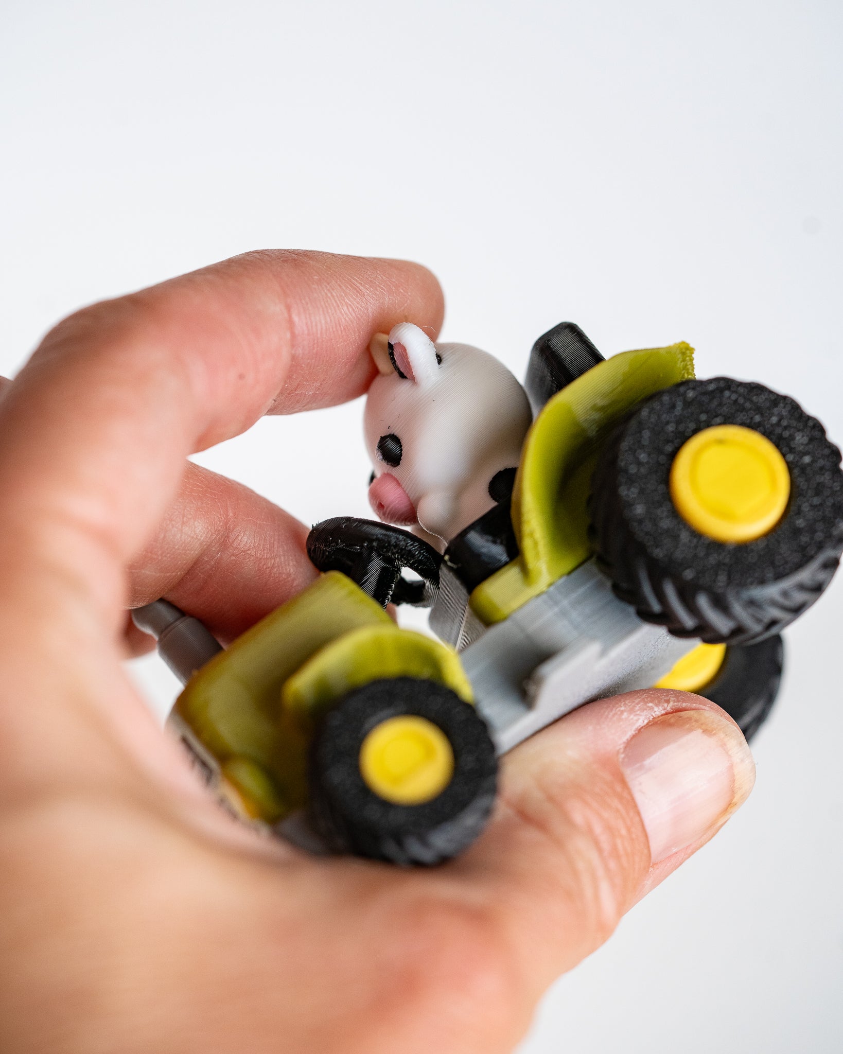A close-up of a hand holding a small toy tractor with a white cow character driving it, photographed against a clean white background.