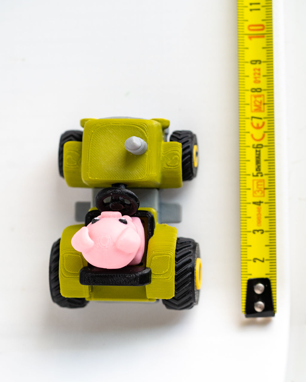 A small yellow toy vehicle with black wheels and a pink pig character in the driver's seat, shown next to a yellow measuring tape on a white background.
