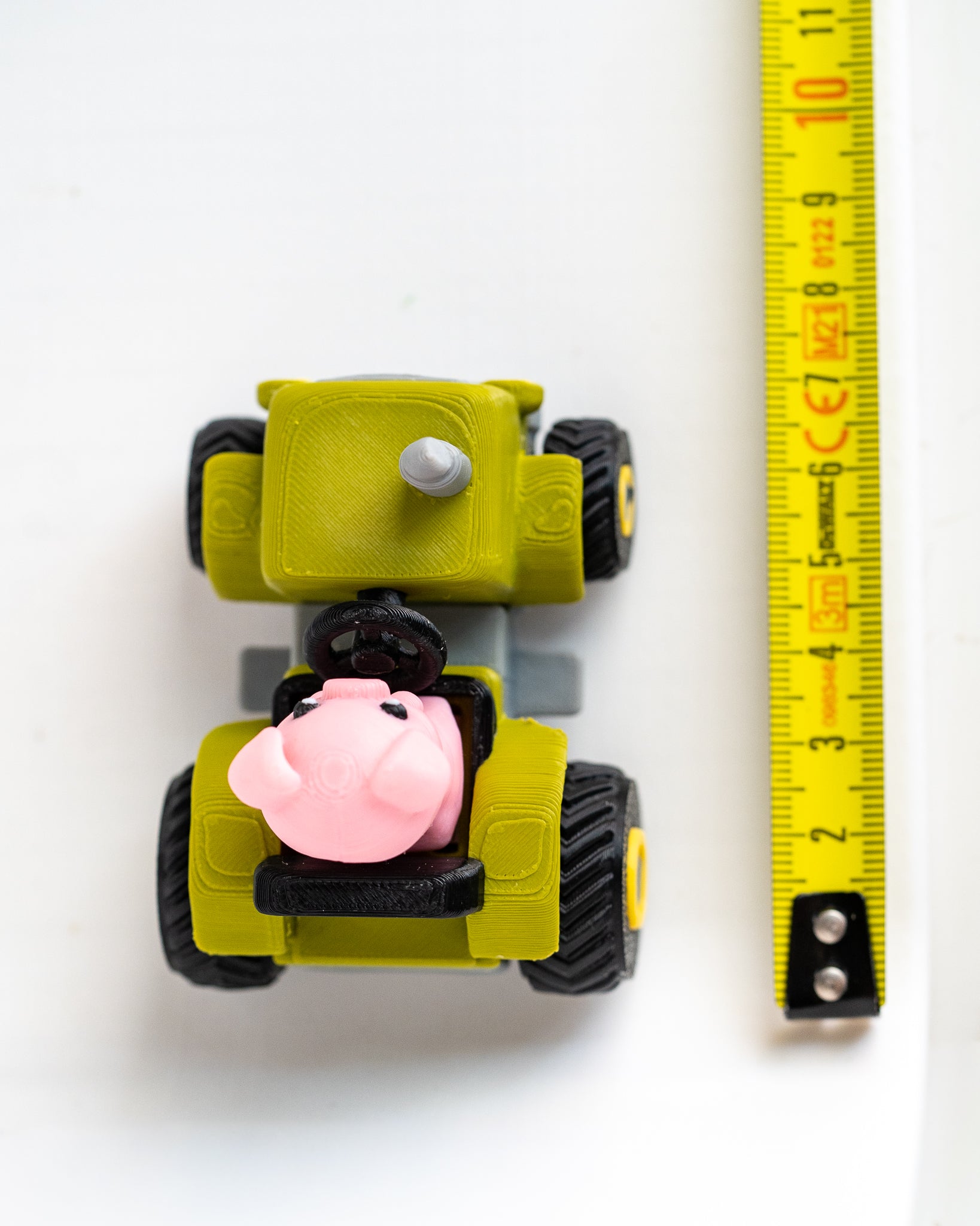 A small yellow toy vehicle with black wheels and a pink pig character in the driver's seat, shown next to a yellow measuring tape on a white background.