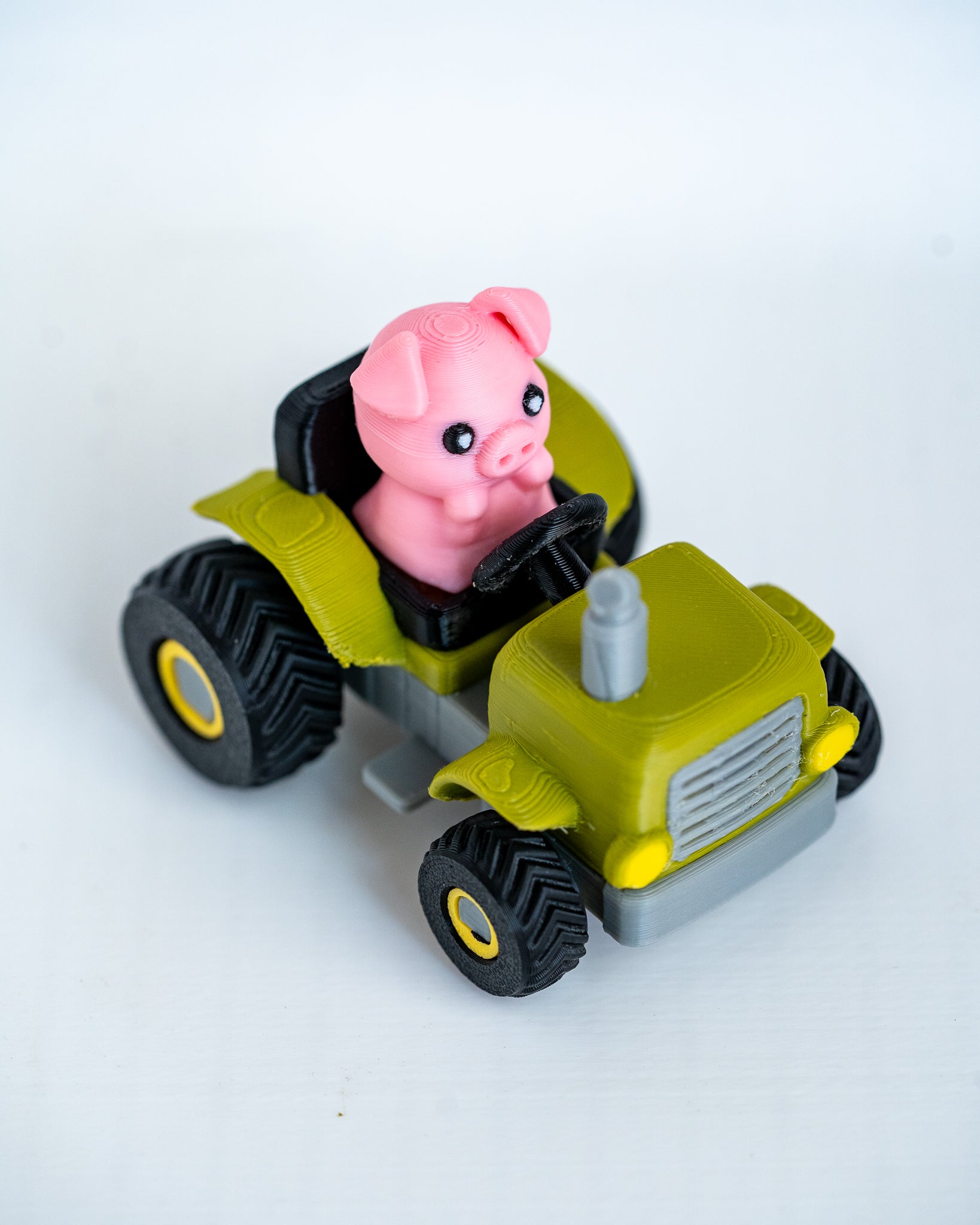 A small toy tractor in olive green, black, and gray with a pink pig figure sitting in the driver's seat, photographed on a plain white background.