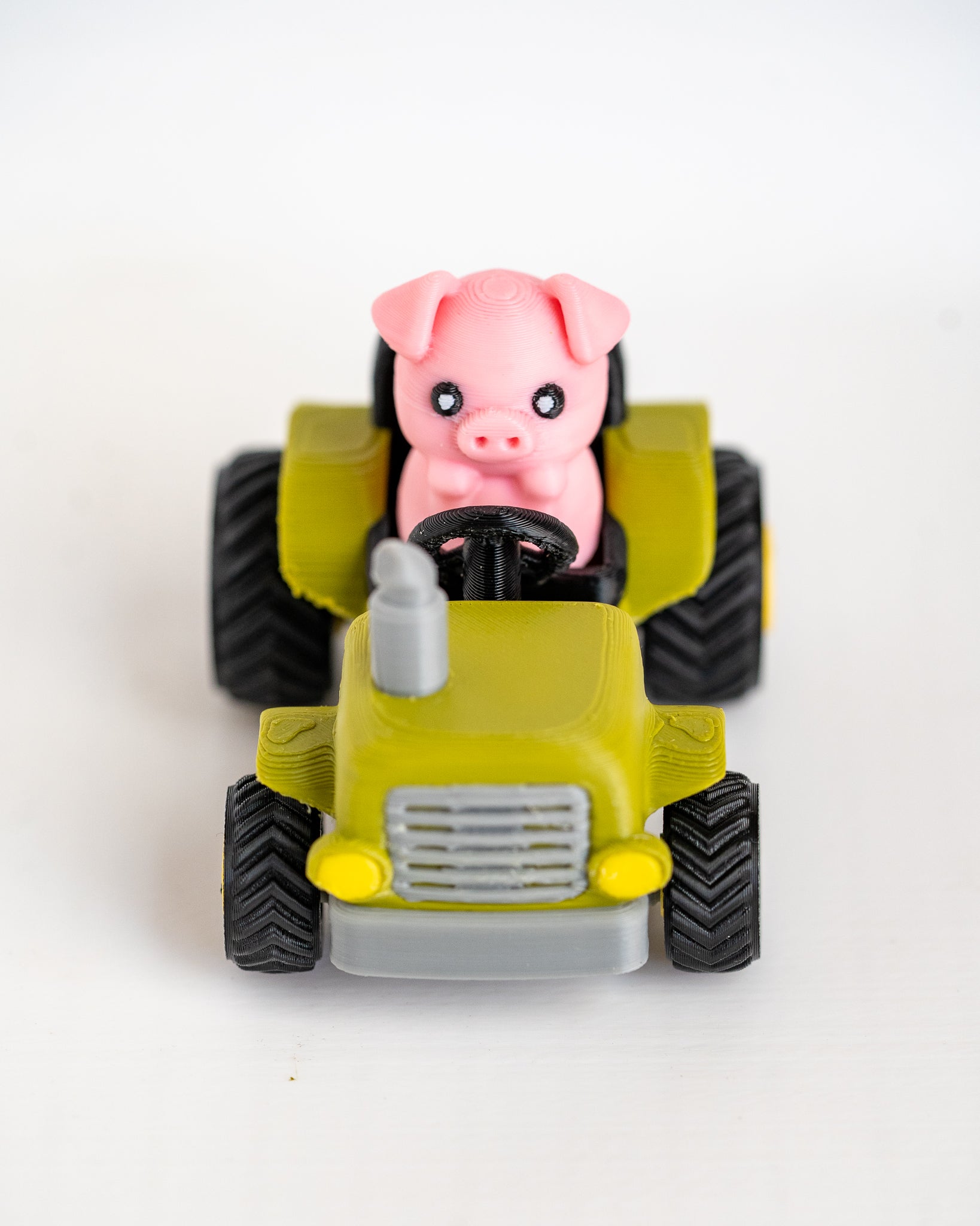 A small toy tractor in yellow-green with black wheels, featuring a pink pig figure sitting in the driver's seat, photographed on a plain white background.