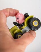 A close-up of a hand holding a small toy vehicle with a pink pig driver. The toy has a lime green body, black wheels with yellow centers, and a gray steering wheel.