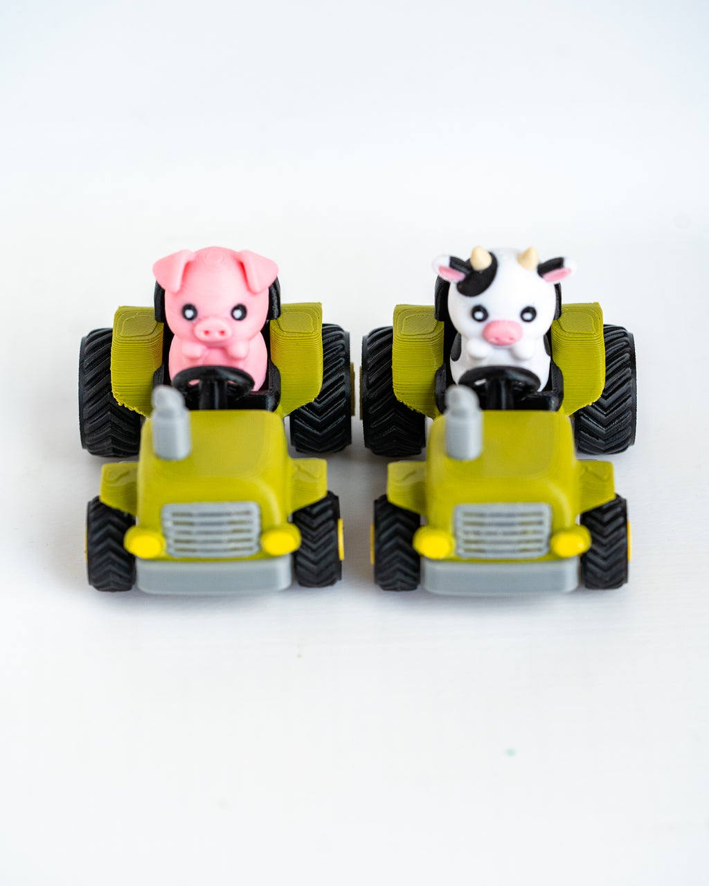Two small toy farm vehicles on a white background: a pink pig driving a green tractor and a black-and-white cow driving another green tractor.