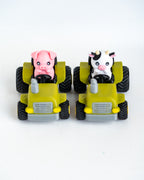 Two small toy farm vehicles on a white background: a pink pig driving a green tractor and a black-and-white cow driving another green tractor.
