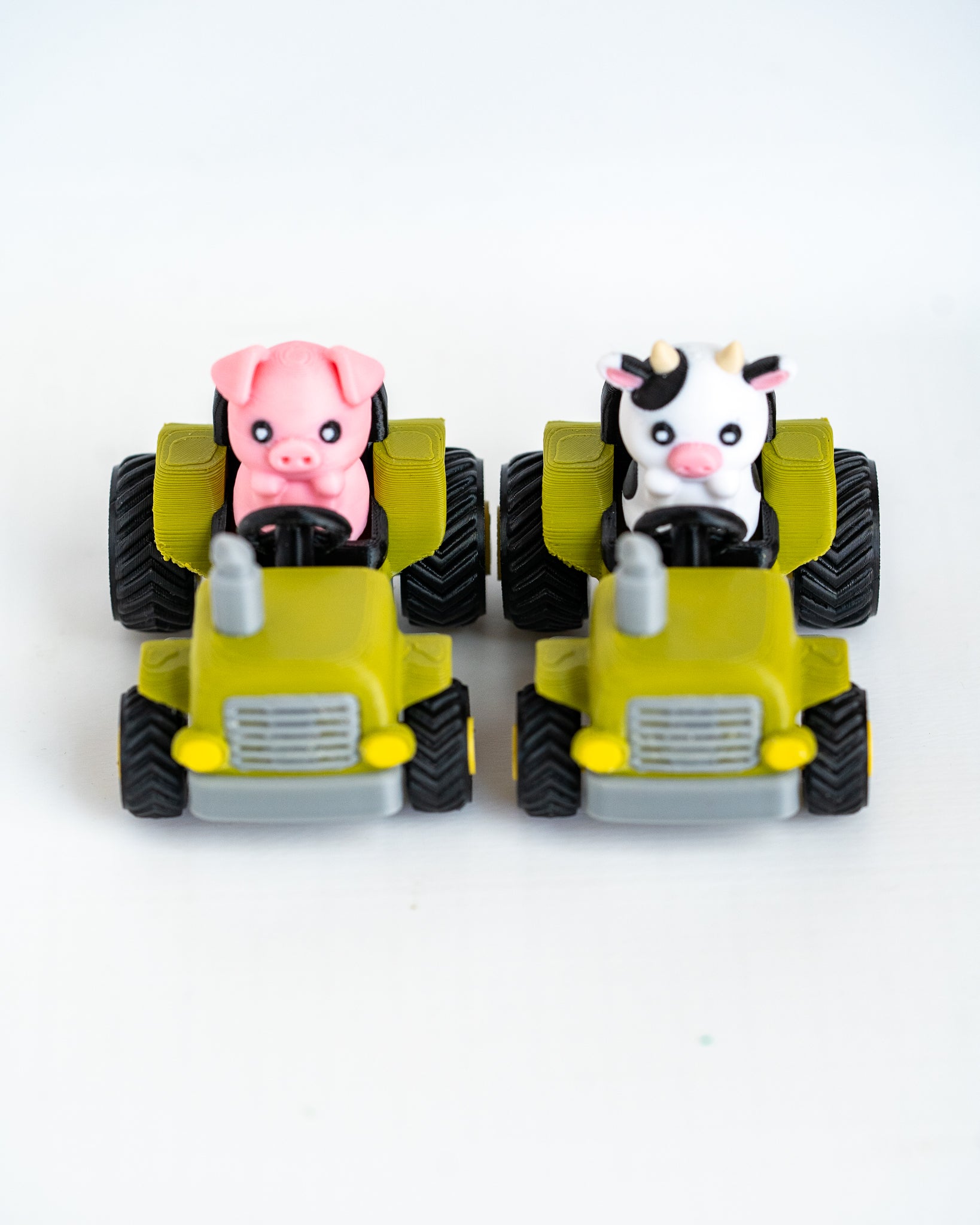Two small toy farm vehicles on a white background: a pink pig driving a green tractor and a black-and-white cow driving another green tractor.