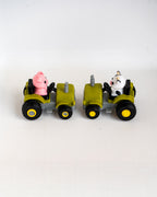 Two small toy tractors on a plain white background. The left tractor is olive green with black wheels and a pink pig character driving it.