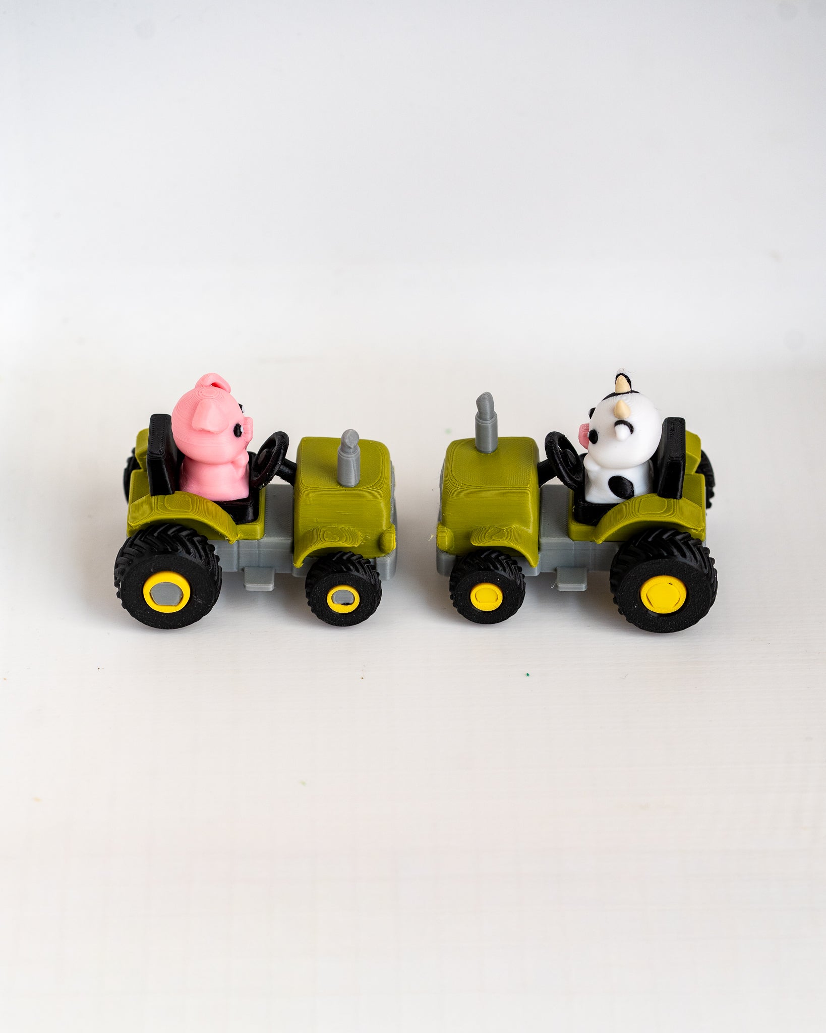 Two small toy tractors on a plain white background. The left tractor is olive green with black wheels and a pink pig character driving it.