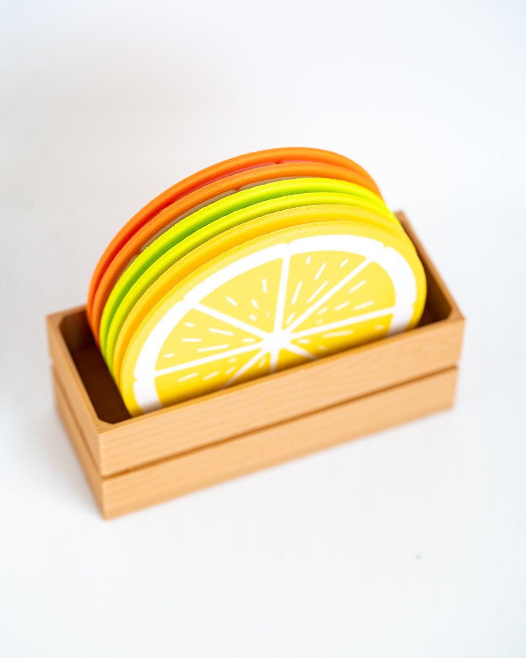 A set of colorful wooden coasters stacked inside a light natural wood box, featuring citrus designs including a yellow lemon slice and orange and green circular pieces, on a white background.