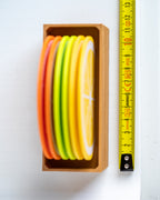 A set of colorful citrus slice coasters in a wooden tray, shown next to a yellow tape measure on a white background.