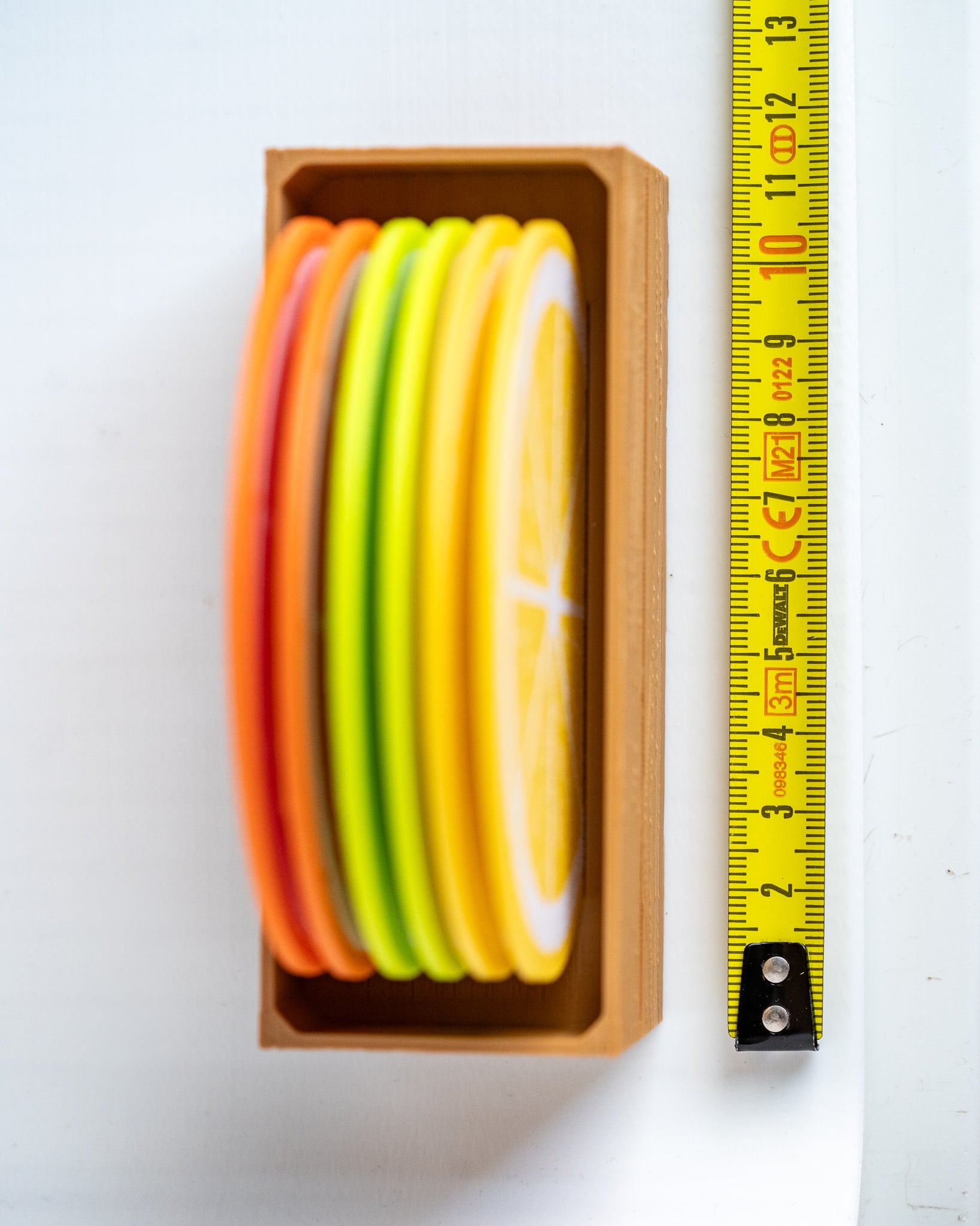 A set of colorful citrus slice coasters in a wooden tray, shown next to a yellow tape measure on a white background.