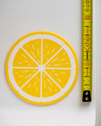 A round yellow and white citrus slice design, likely a coaster or patch, shown next to a yellow measuring tape on a white background.