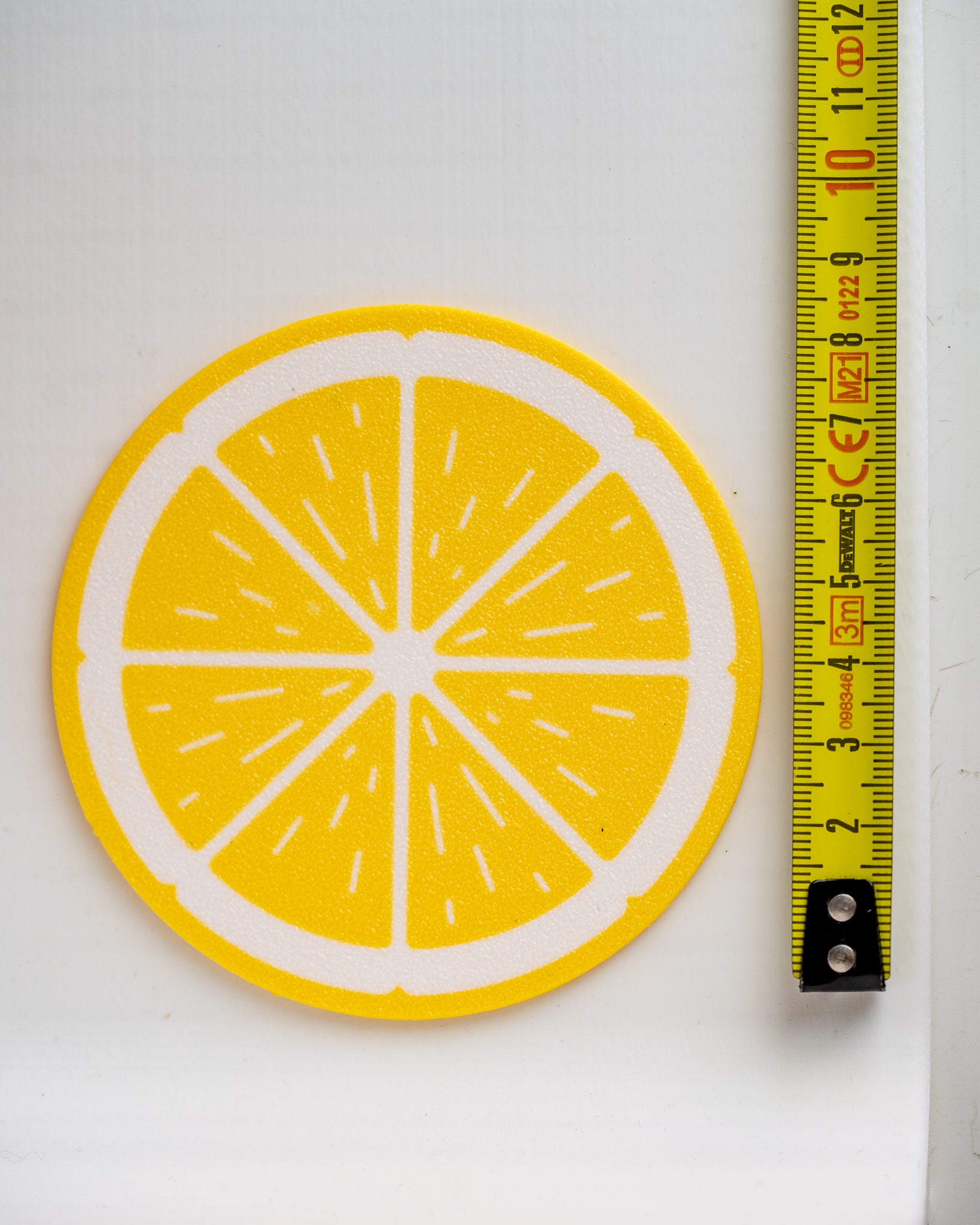 A round yellow and white citrus slice design, likely a coaster or patch, shown next to a yellow measuring tape on a white background.