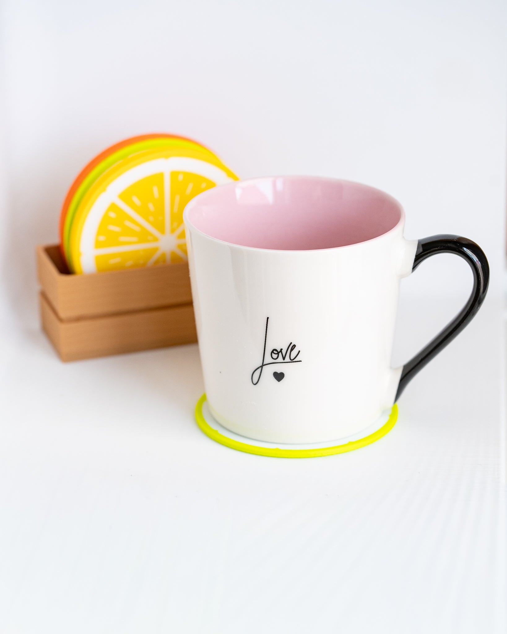 A white ceramic coffee mug with a pink interior, black handle, and black 'love' script on the front, sitting on a neon yellow coaster. A stack of colorful citrus slice coasters rests in a small wooden holder in the background against a white studio backdrop.