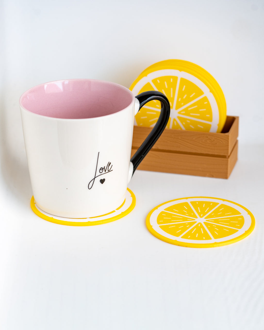 A white ceramic mug with a pink interior and black handle sits on a yellow lemon-shaped coaster. A matching lemon coaster lies in front, and a wooden box holding additional lemon coasters is behind the mug.