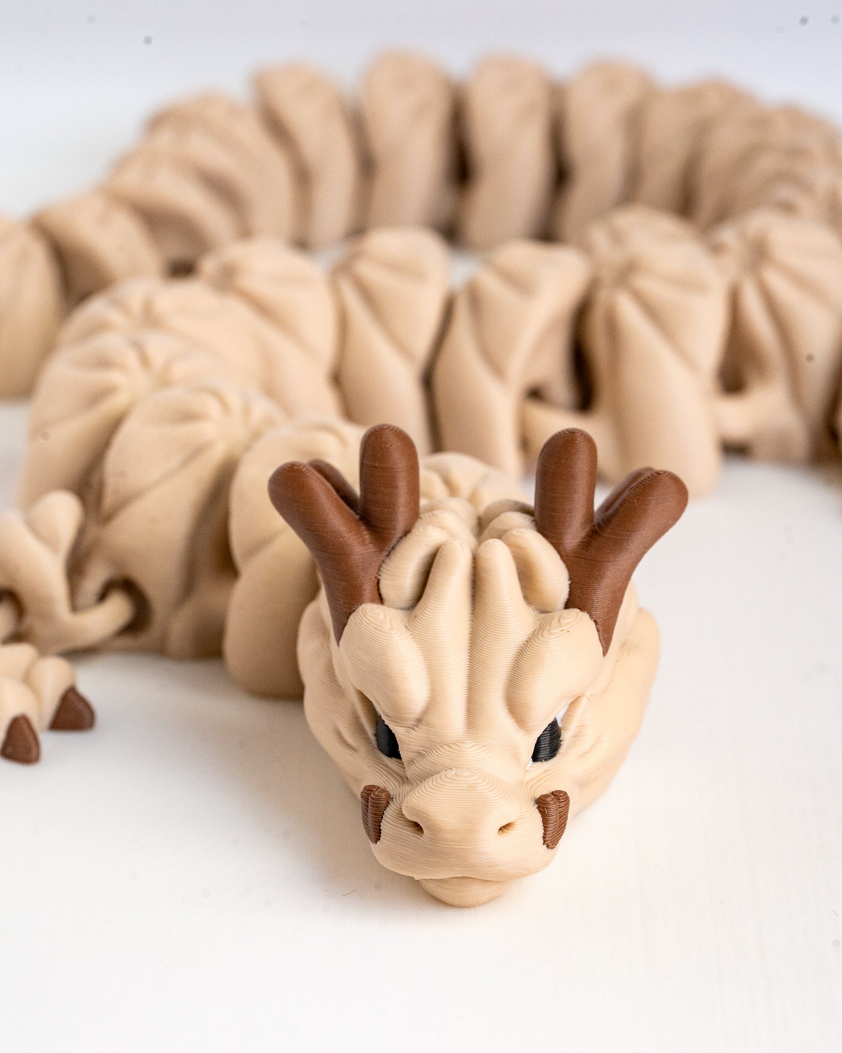 A beige 3d printed dragon figurine with brown horns and black eyes, shown on a white surface with its body blurred in the background.