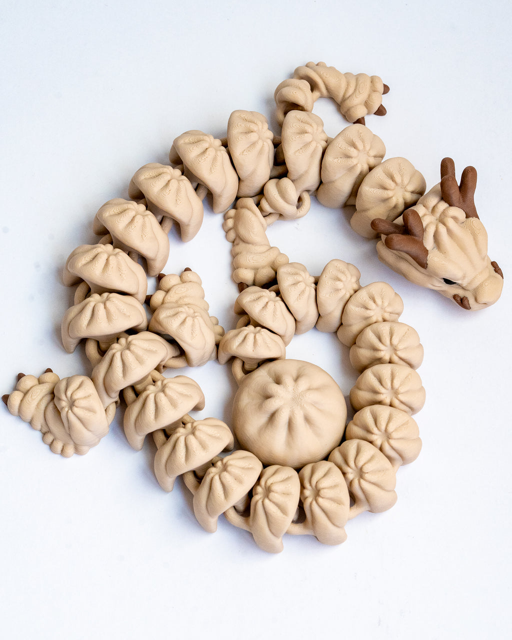 A 3d printed dragon made from small dumpling on a plain white background. The dragon has a light beige body with darker brown antlers and facial details.