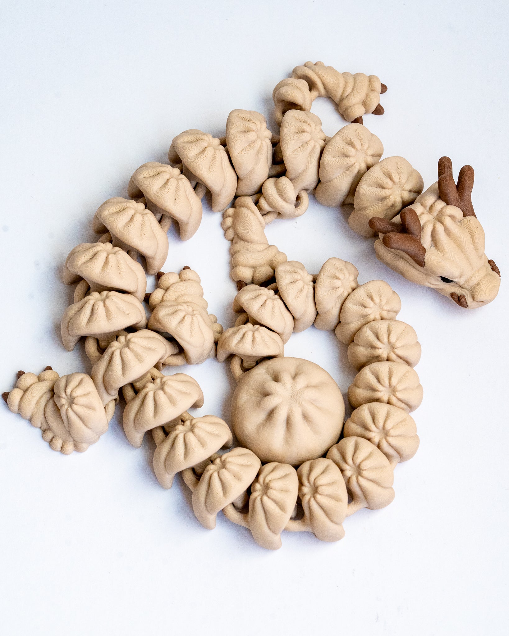 A 3d printed dragon made from small dumpling on a plain white background. The dragon has a light beige body with darker brown antlers and facial details.