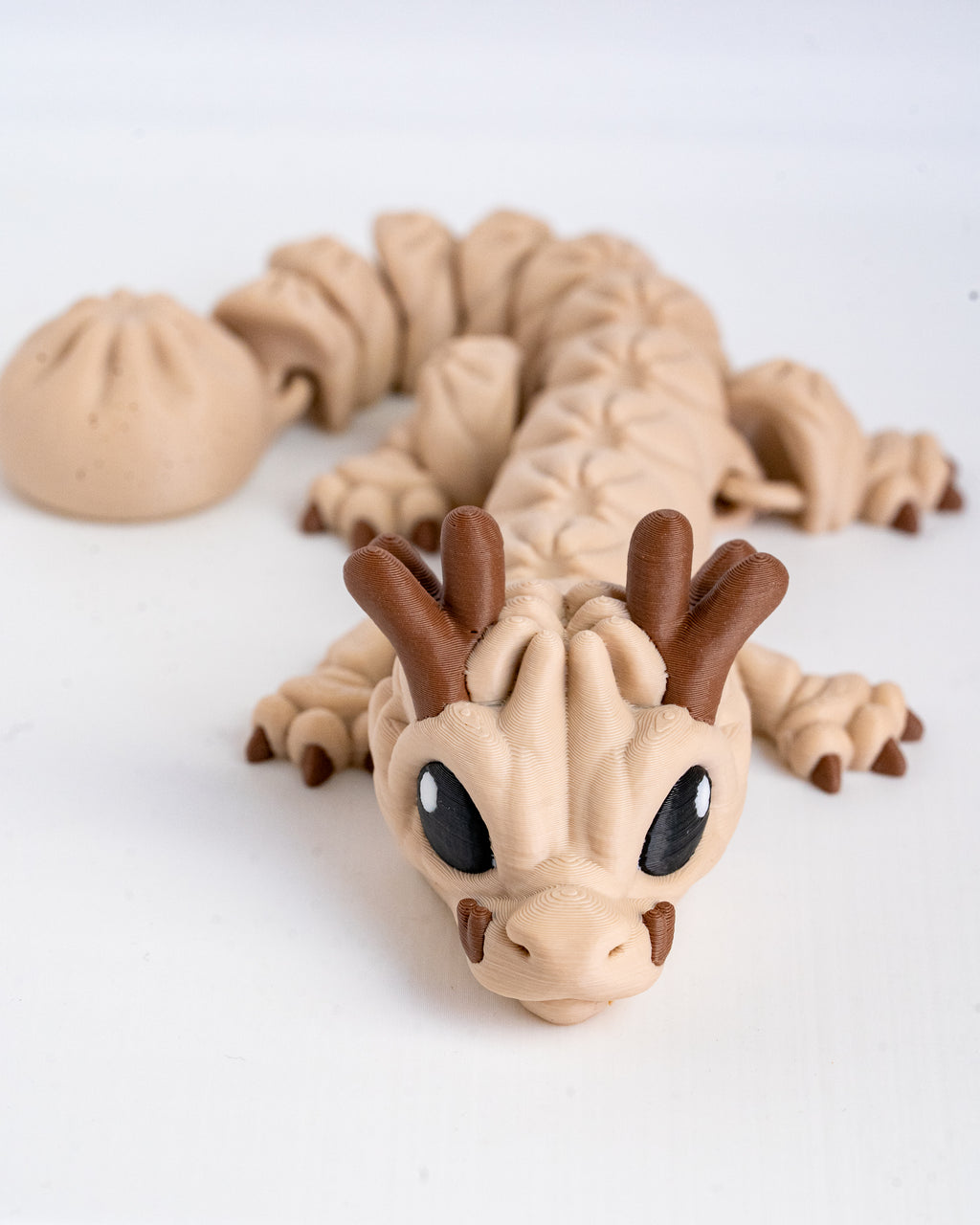 A small 3d printed dragon figurine with a beige body, brown horns and claws, and large black eyes, photographed on a plain white background.