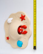 A beige sandcastle-shaped toy with a red crab peeking out from the center, plus small red starfish, a white shell, and a blue shell detail, shown on a white background next to a yellow measuring tape.