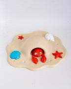 A children's beach-themed play mat featuring a beige sand-like base with a red lobster peeking out from a shell-shaped opening, surrounded by blue and red seashells and a white cloud detail, photographed on a white background.