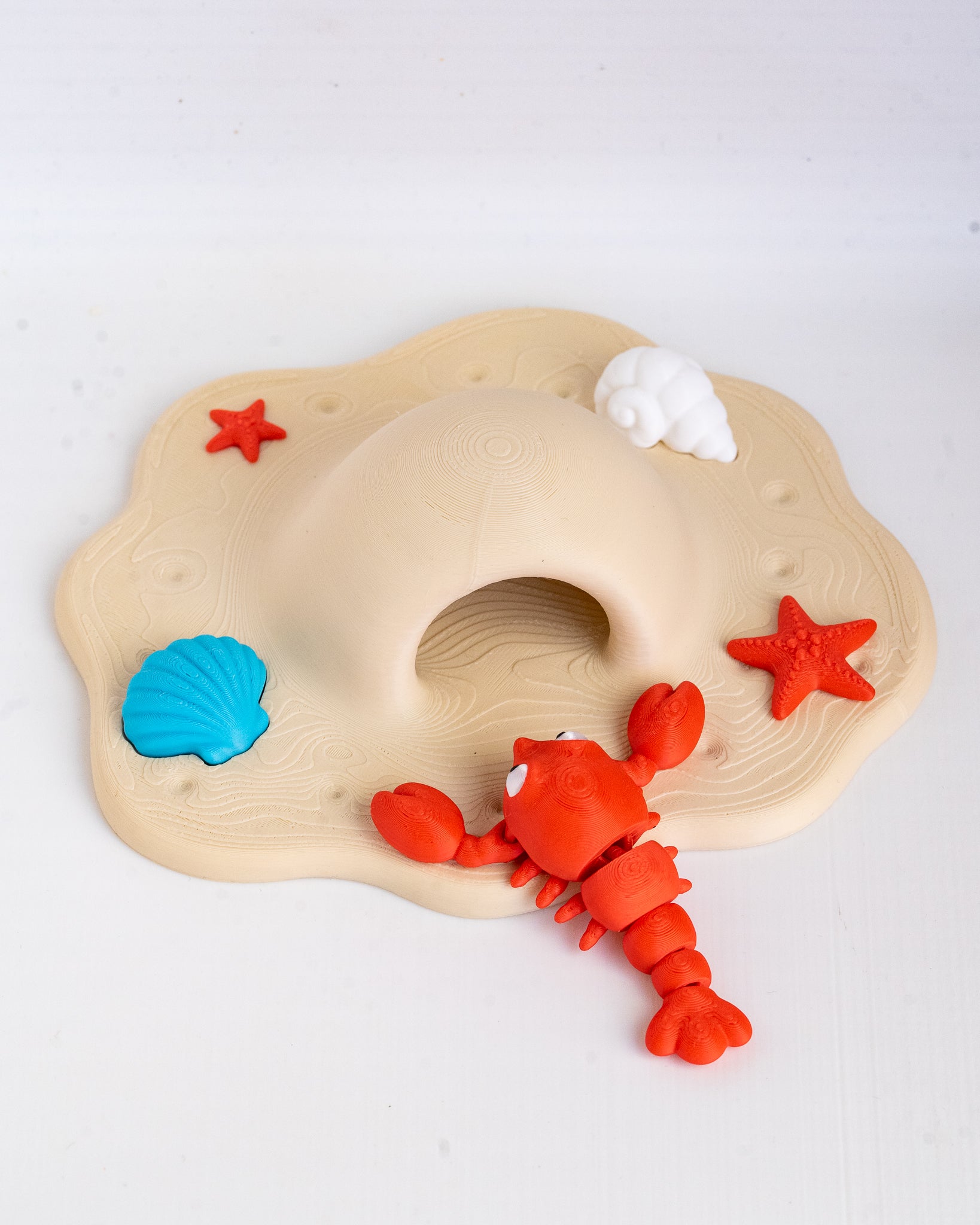 A children's toy beach scene with a beige sandcastle base, red lobster, blue shell, white shell, and red starfish on a white background.