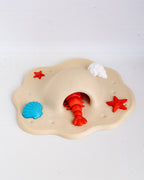 A children's beach-themed sensory toy with a beige sand mound, a red lobster hole and lobster, a blue shell, a white shell, and red starfish on a white background.