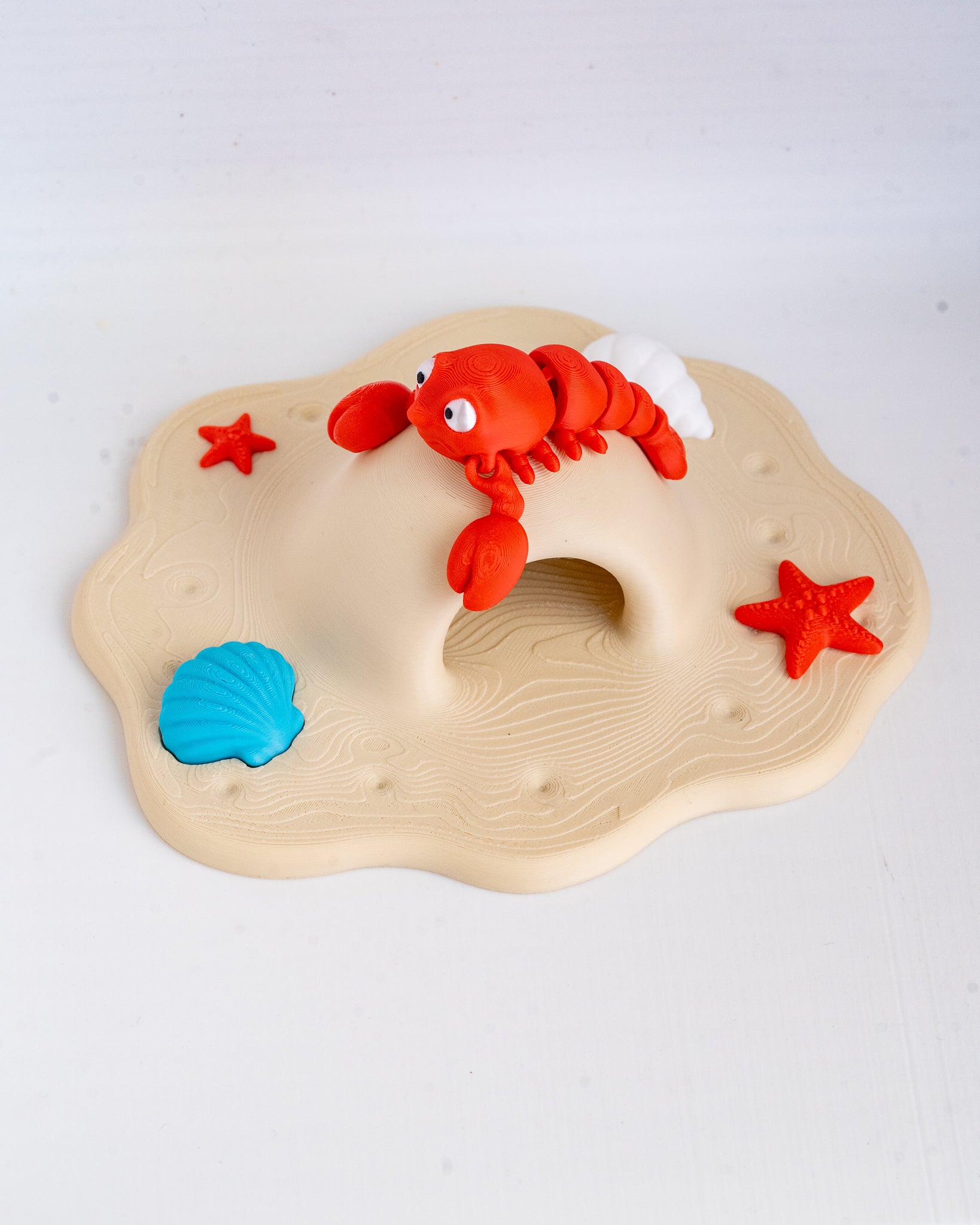 A children's toy sand play set featuring a beige sand base with a molded hole, a red lobster figure, a blue seashell, and two red starfish on a white background.